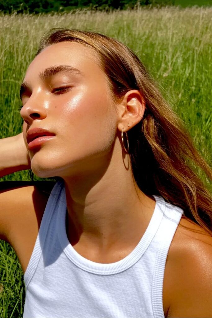 A woman sitting in tall green grass with eyes closed, wearing a white tank top and hoop earrings, photographed in bright natural sunlight with a calm, relaxed expression.